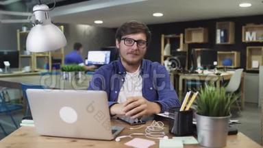 幸福企业主的肖像，好看的胡须男，戴着眼镜，坐在创意办公室，看着相机。